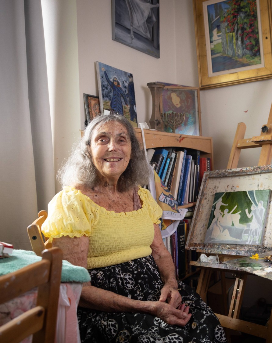 Older woman sitting inside her home with paints and painting surrounding her