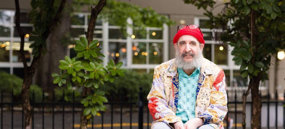Older man wearing a sweater sitting on a bench outside smiling at the camera