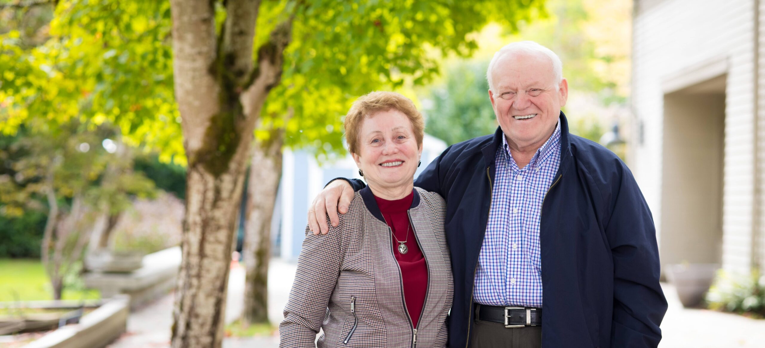 Older husband and wife standing smiling for a photo. Husband's arm around the wife.