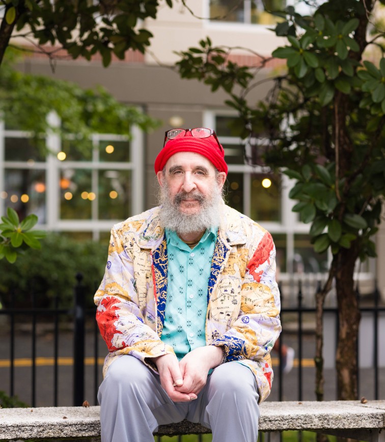 Older man wearing a sweater sitting on a bench outside smiling at the camera