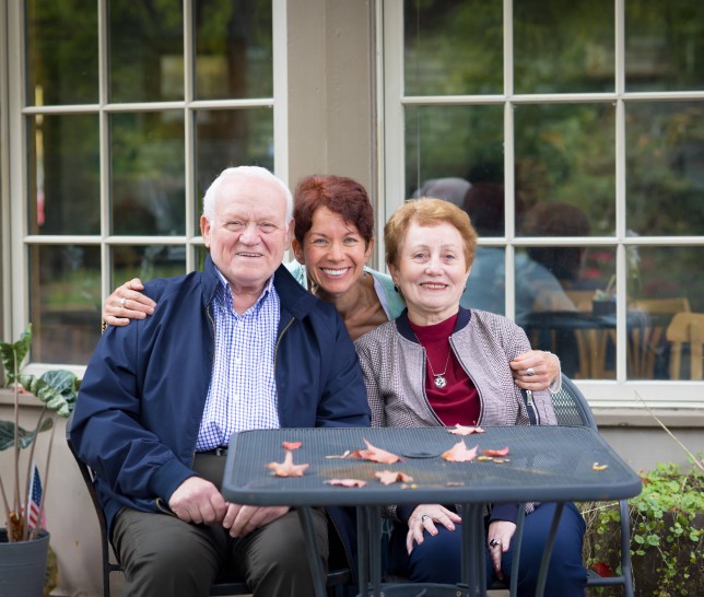 An elderly couple sitting at a table smiling with a nurse squating between them with her arms around them