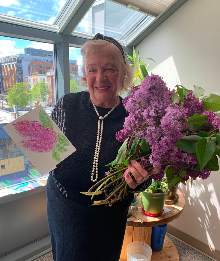 Older woman holding purple flowers and a painted card with purple flowers on it.