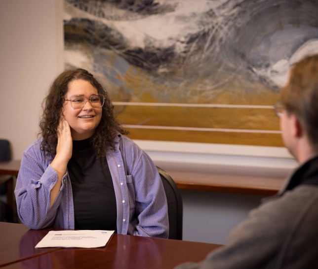 Woman sitting at a conference desk is speaking to a man across from them.