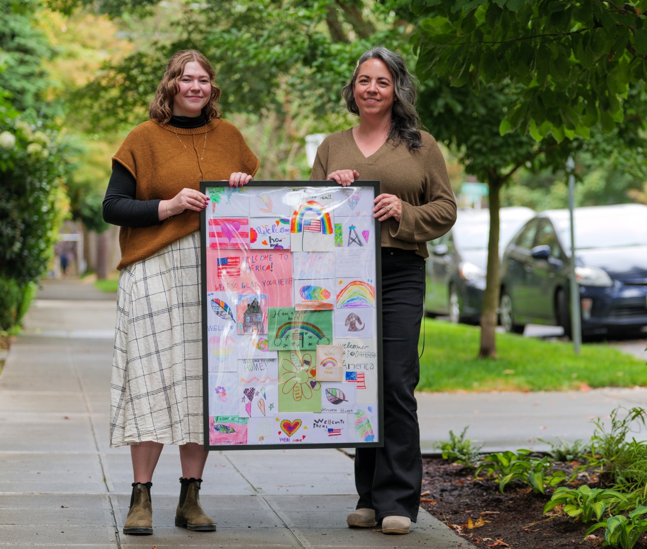 Two women holding a frame filled with cards of drawing from kids in it