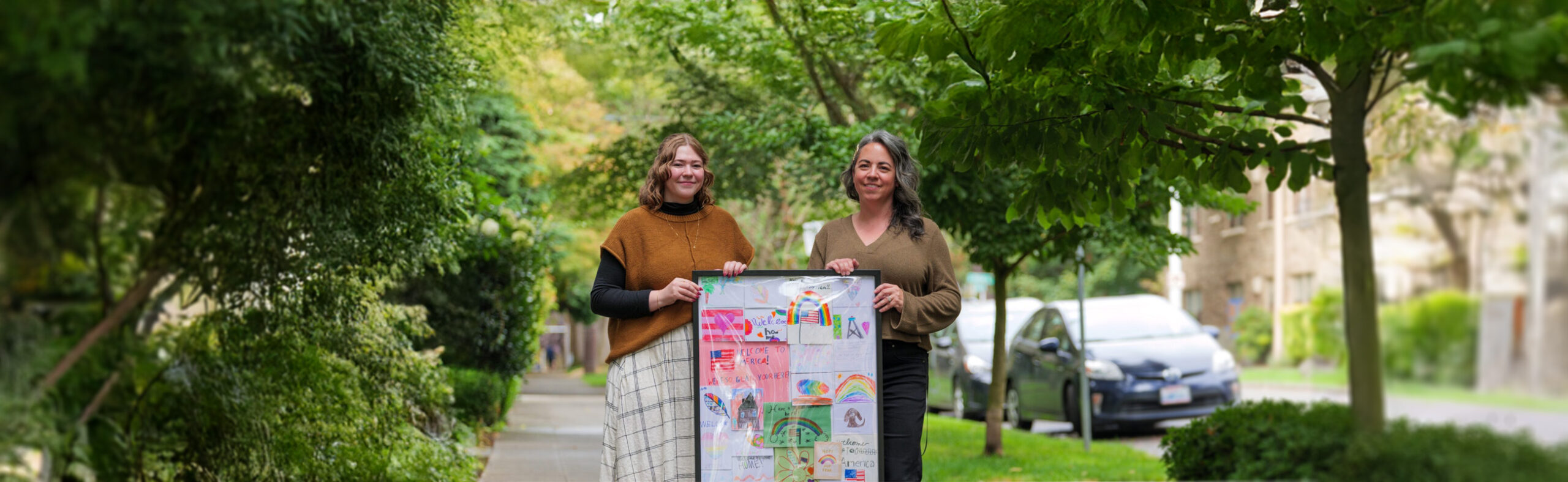 Two women holding a frame filled with cards of drawing from kids in it